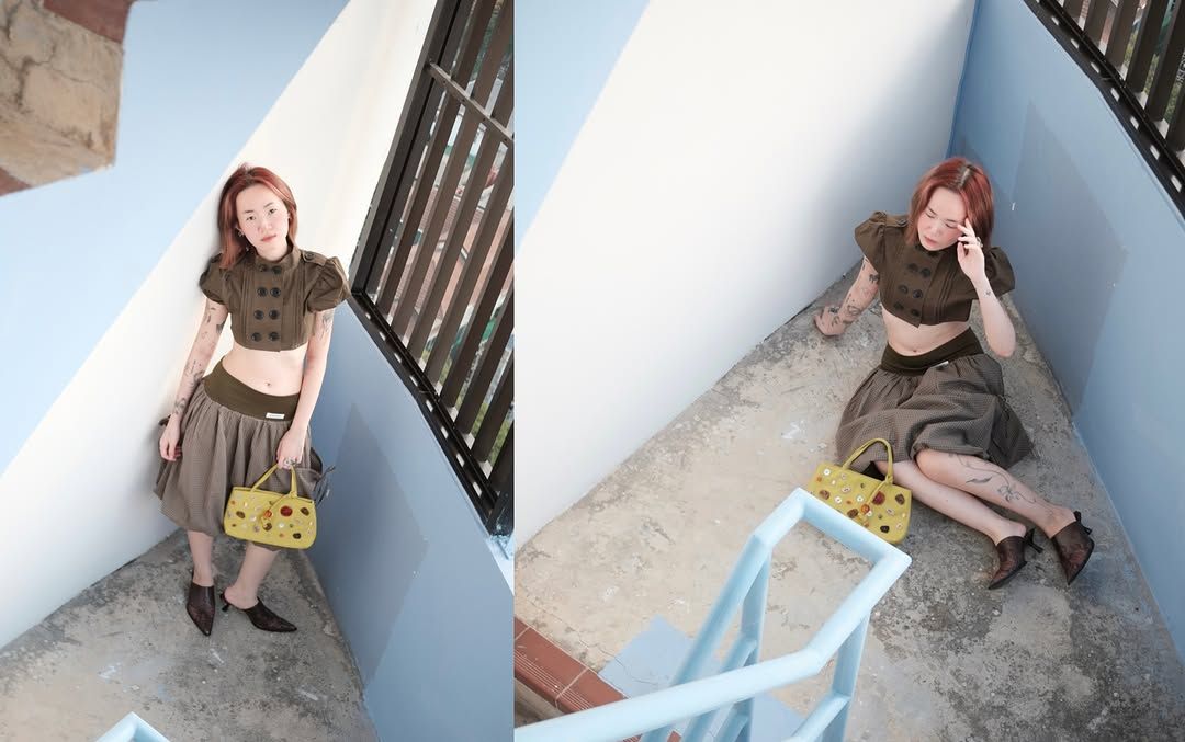A woman with red hair poses in a stylish outfit featuring a cropped top and a long skirt, holding a yellow handbag while sitting in a modern, angular space.