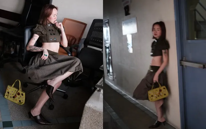 A model poses in a stylish outfit featuring a cropped top and a long skirt, showcasing a unique handbag. The setting has an urban feel with a mix of indoor and outdoor elements.