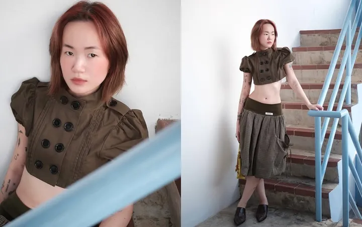 A model poses in a stylish outfit featuring a cropped, button-up top and a long skirt, standing by a blue staircase.