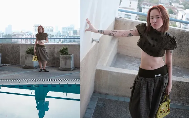 A person stands on a rooftop, wearing a cropped top and a long skirt, with a reflection visible in the pool beside them. The city skyline is seen in the background.