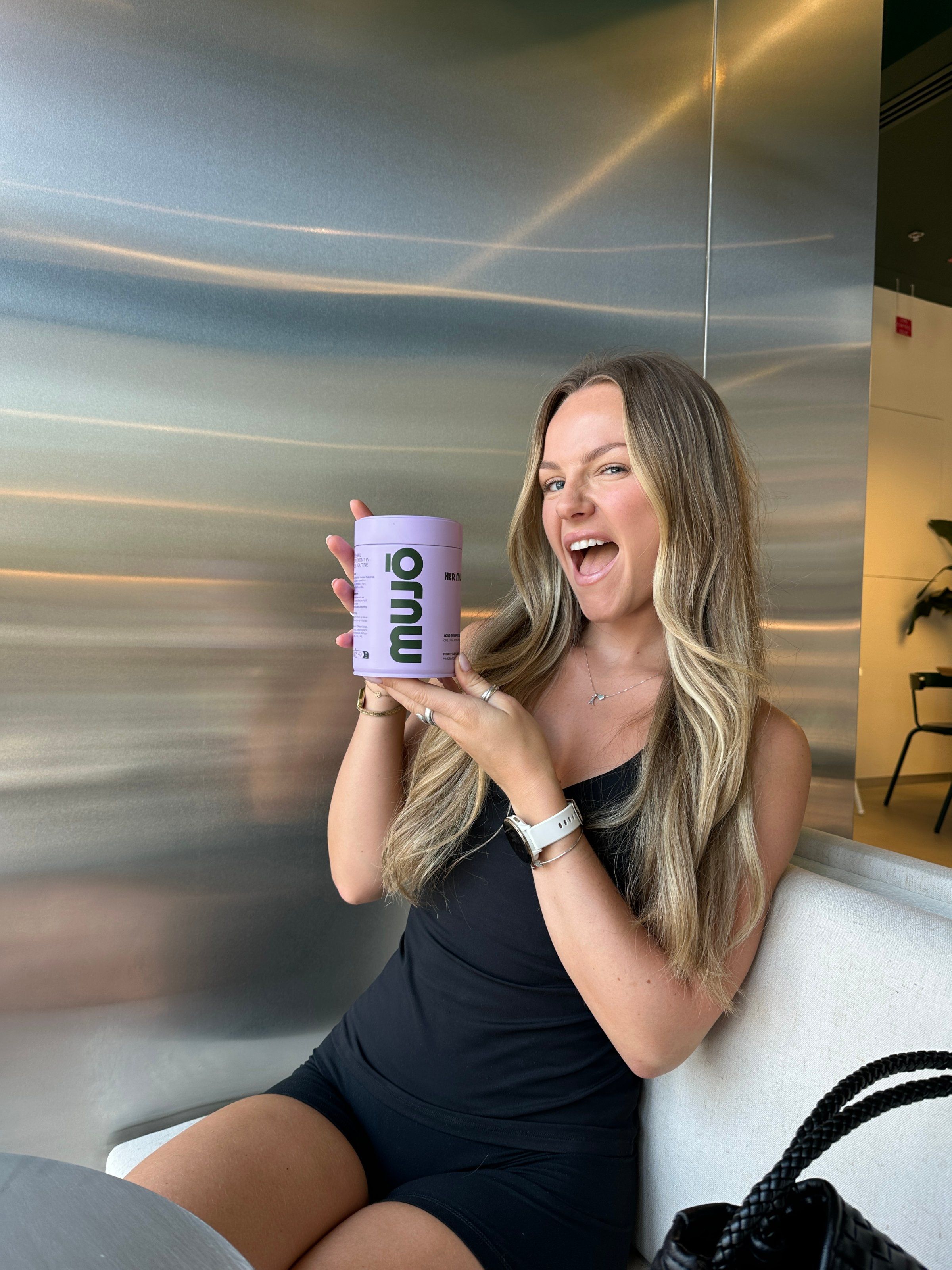 A woman with long hair smiles while holding a purple container labeled 'muu' in a modern indoor setting.