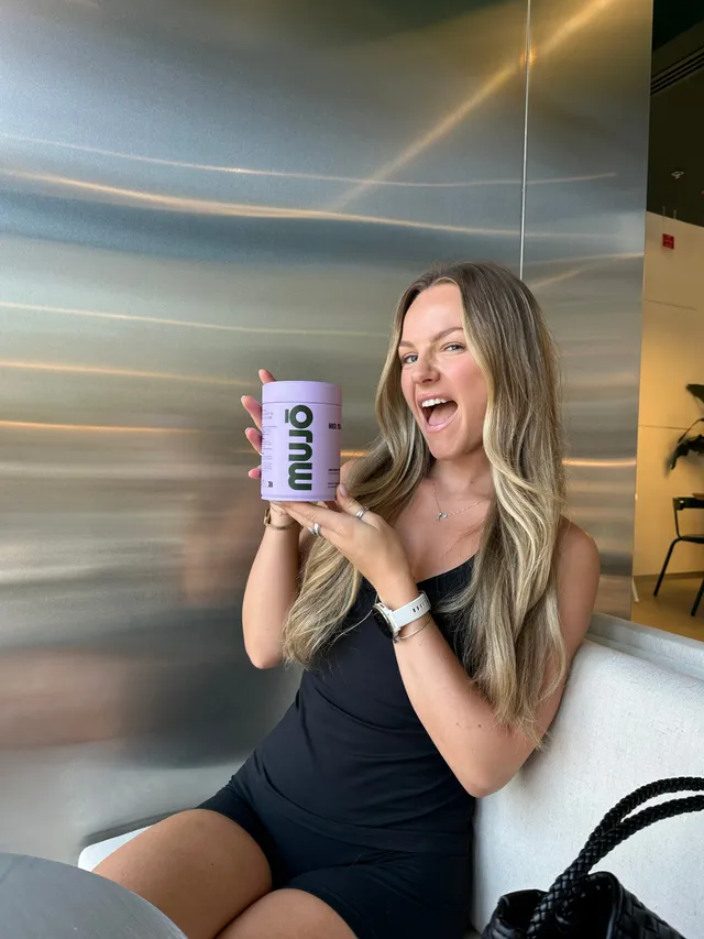 A woman with long hair smiles while holding a purple container labeled 'muu' in a modern indoor setting.
