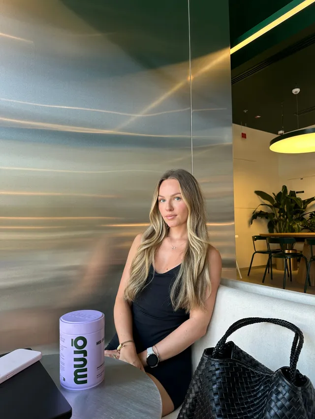 A young woman with long blonde hair sits at a table in a modern café, wearing a black dress and looking confidently at the camera. A purple container labeled 'muwu' is placed on the table beside her.