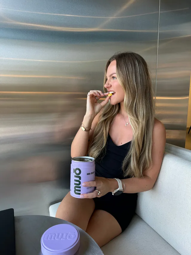 A woman with long hair sitting at a table, enjoying a snack while holding a can of mwū beverage.