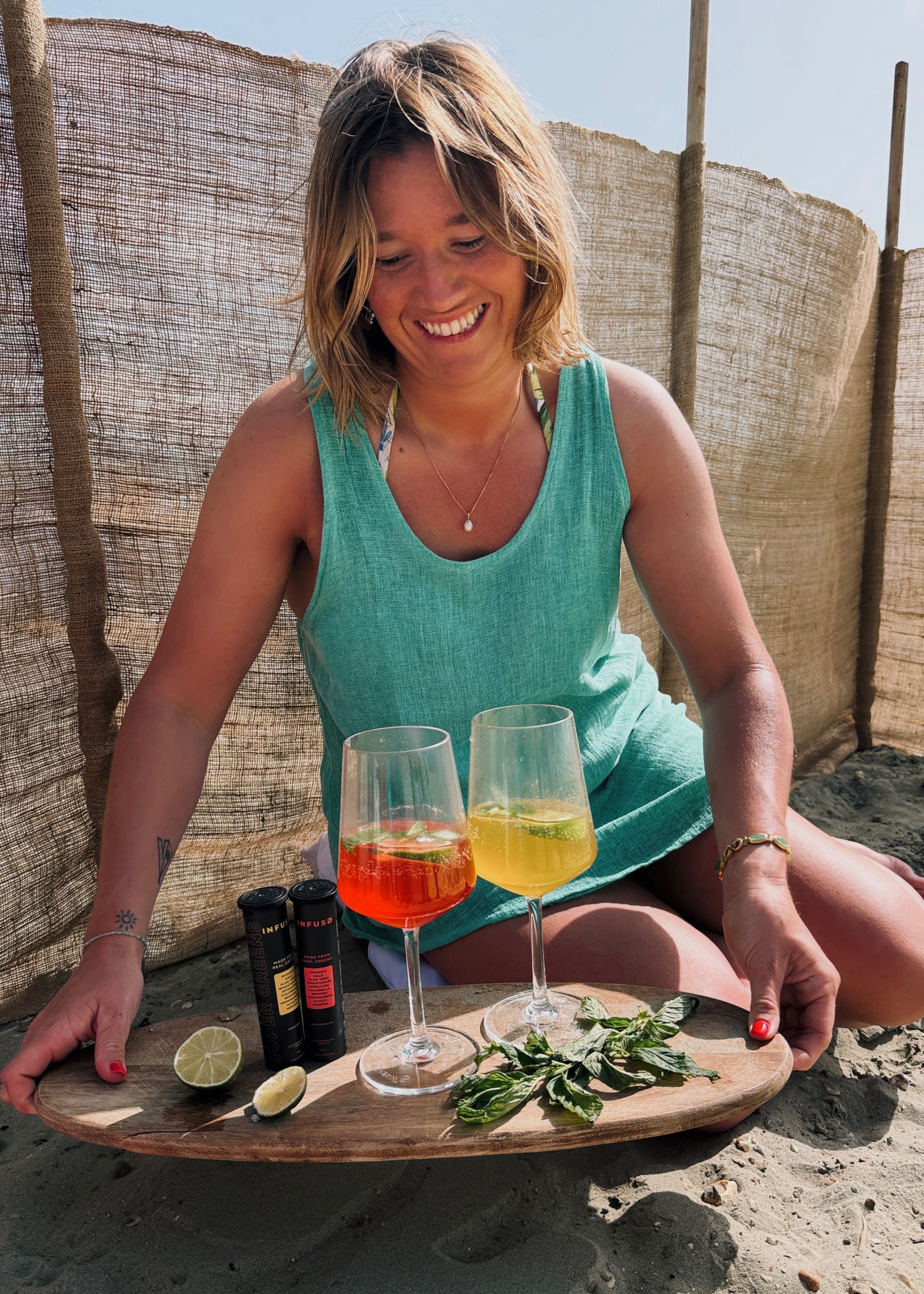 A woman sitting on the sand at the beach, smiling as she presents two colorful cocktails on a wooden platter, surrounded by fresh mint and lime.