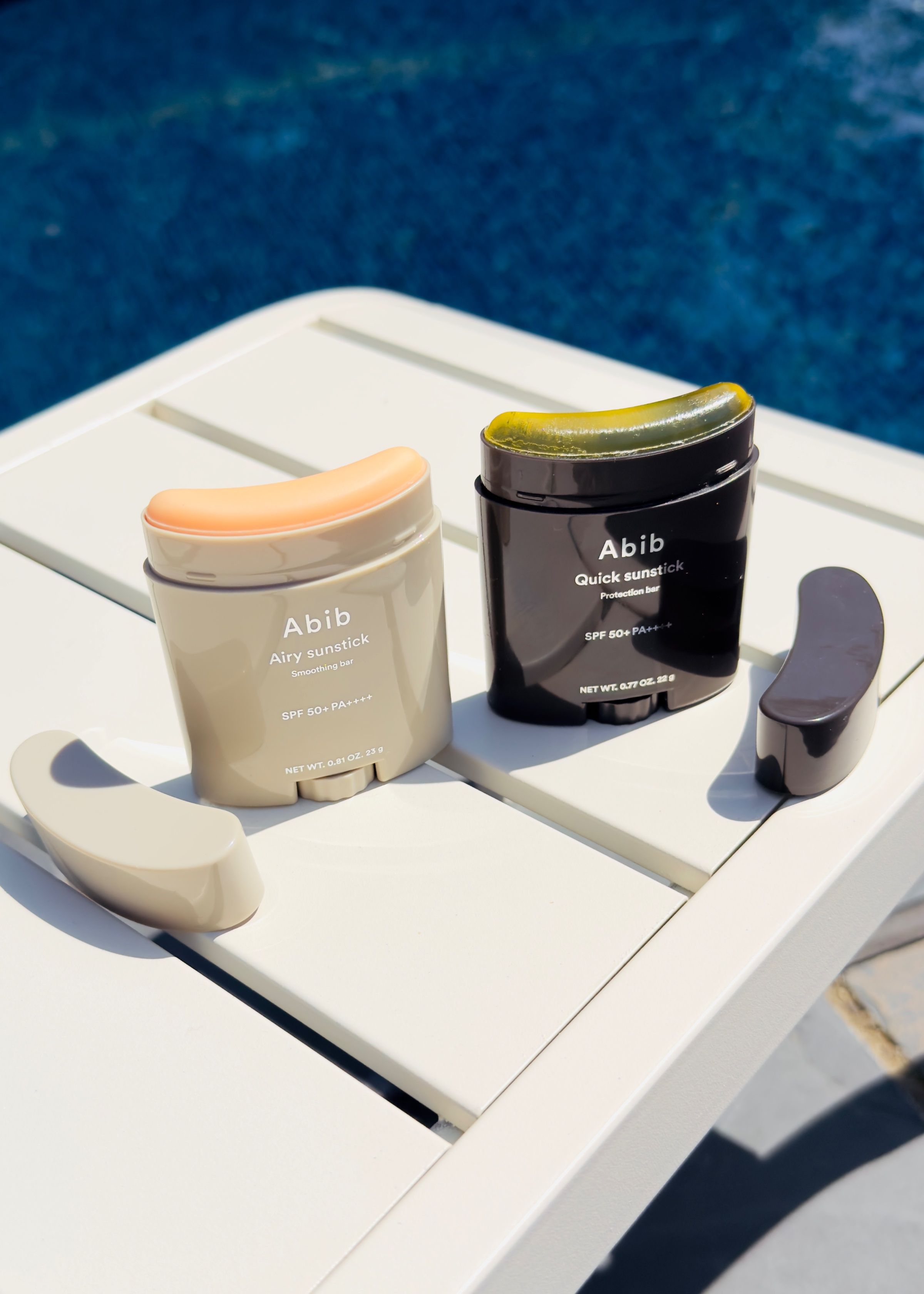 Two skincare products from Abib displayed on a white table beside a swimming pool, showcasing their sleek packaging.
