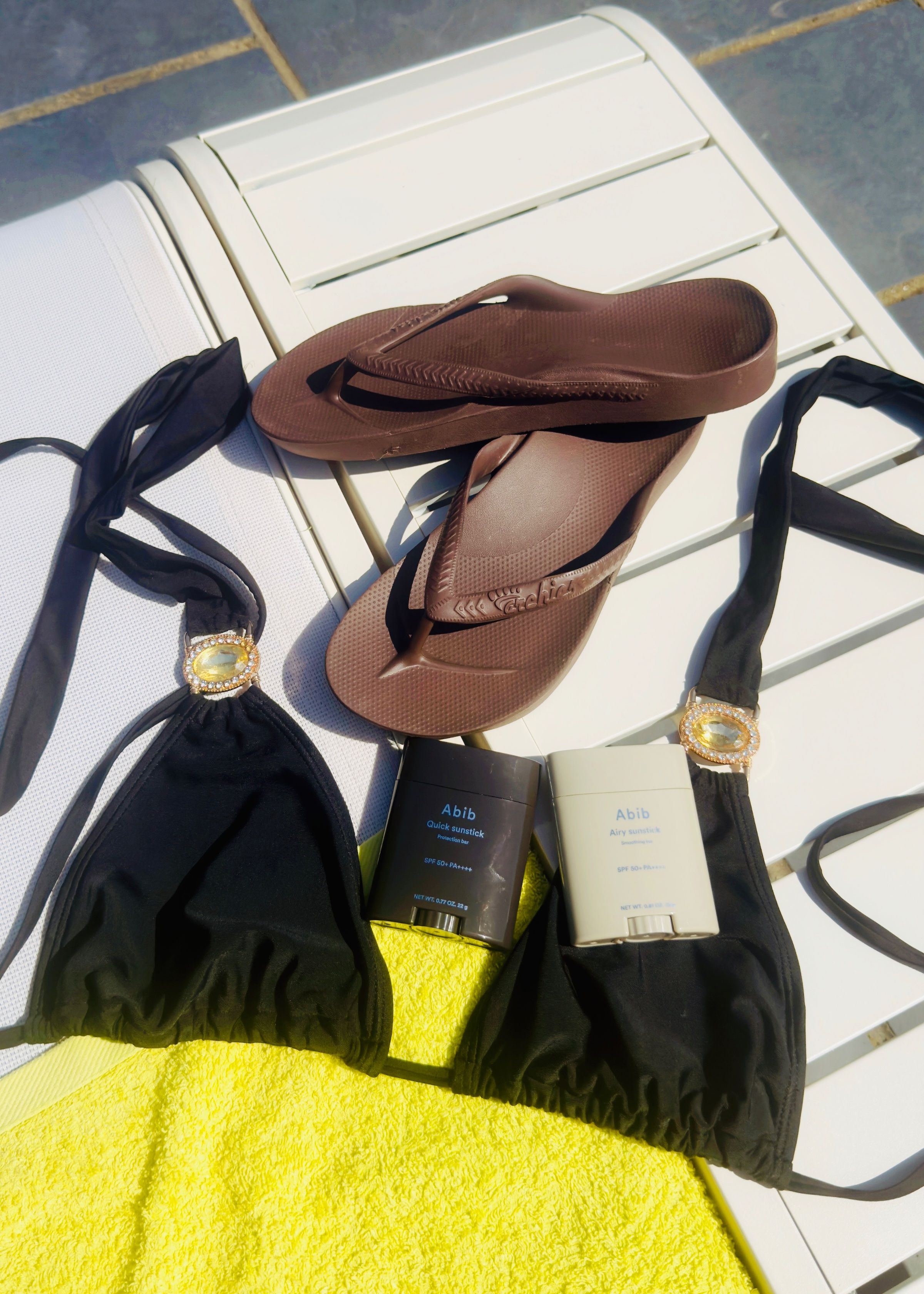 A flat lay of swimwear and accessories on a sun lounger, featuring black bikini tops, a pair of brown flip-flops, and sunscreen products on a bright yellow towel.