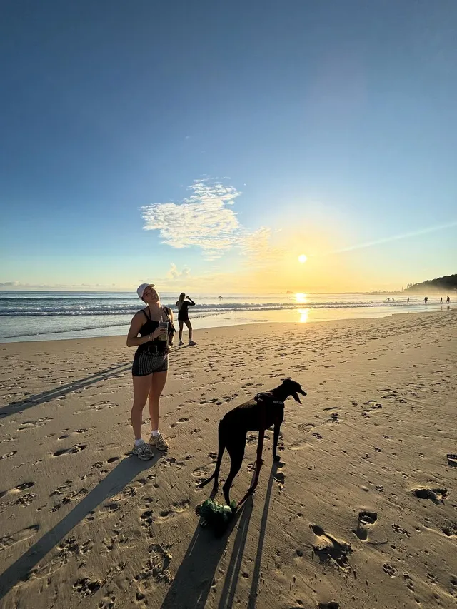 A woman stands on the beach at sunset, looking up with a joyful expression, accompanied by a dog. In the background, other people are enjoying the beach.