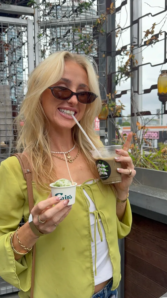 A smiling woman with long blonde hair holds a small cup of green ice cream in one hand and a drink with a straw in the other, wearing stylish sunglasses and a green blouse.
