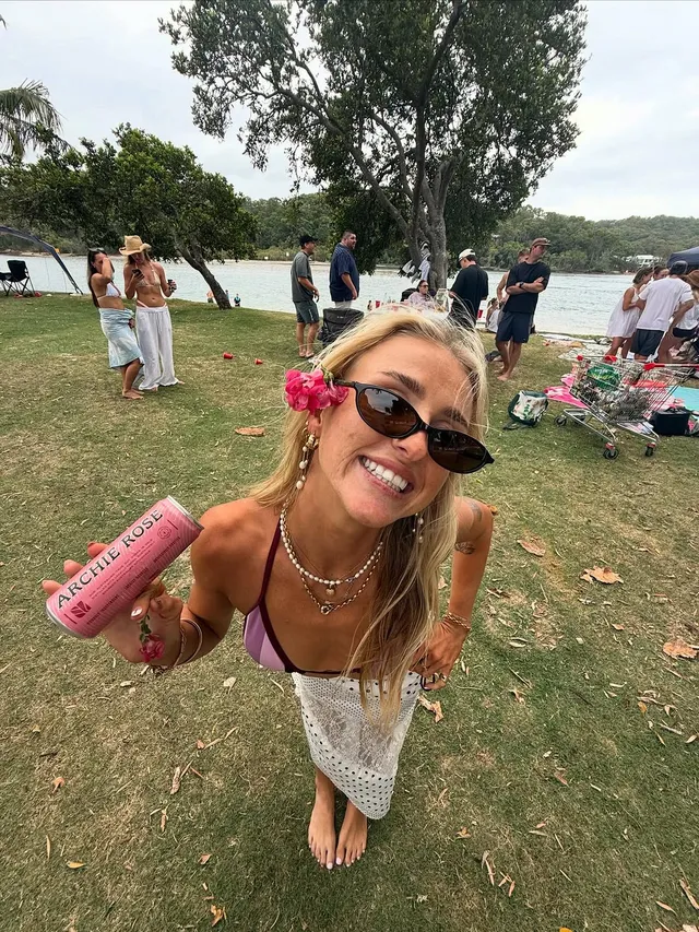 A smiling woman with sunglasses and a flower in her hair holds a drink while posing in a grassy area near a body of water, surrounded by people enjoying a gathering.