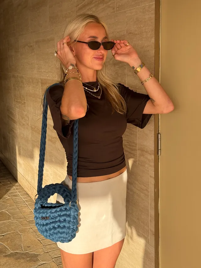 A woman wearing sunglasses and a black top poses with a blue woven handbag against a textured wall.