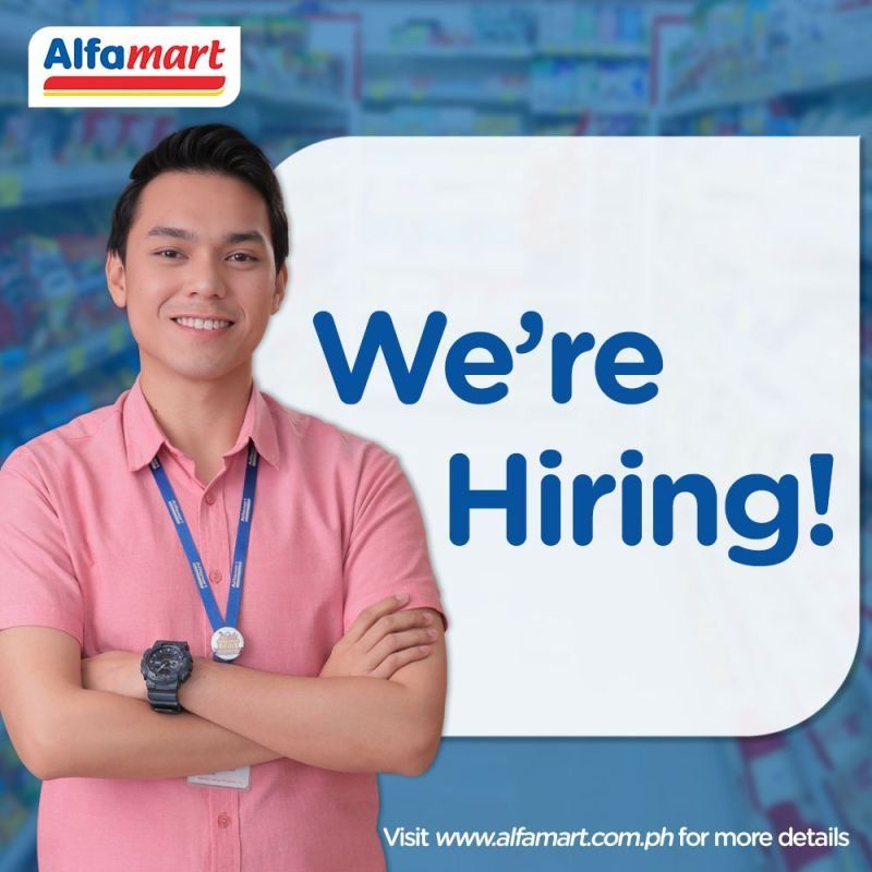 A young man stands confidently with his arms crossed, smiling in a retail environment. The background features shelves stocked with products, and a sign indicating that the store is hiring.