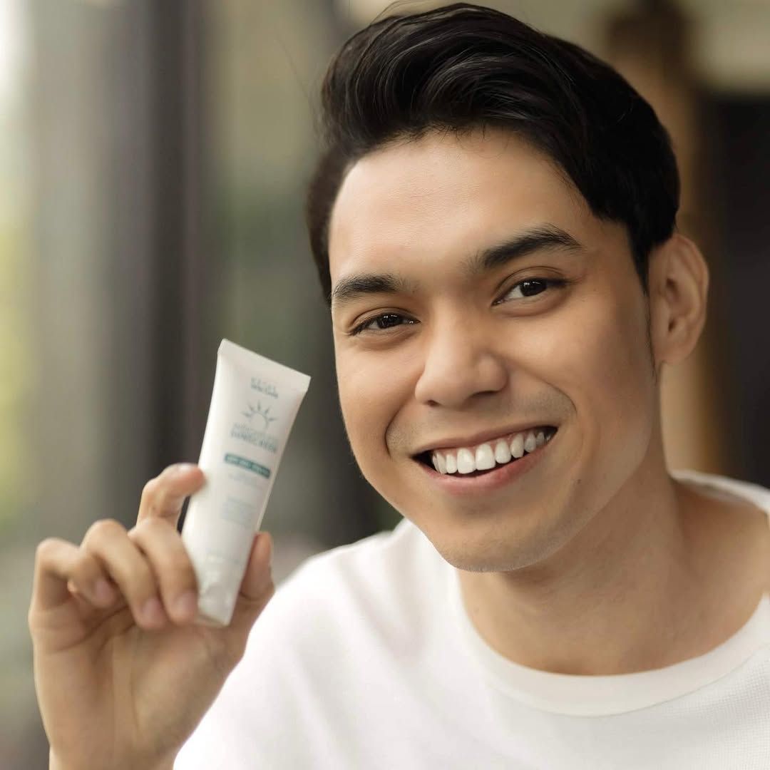 A smiling man holds a skincare product in his hand, showcasing it to the camera with a bright and inviting expression.