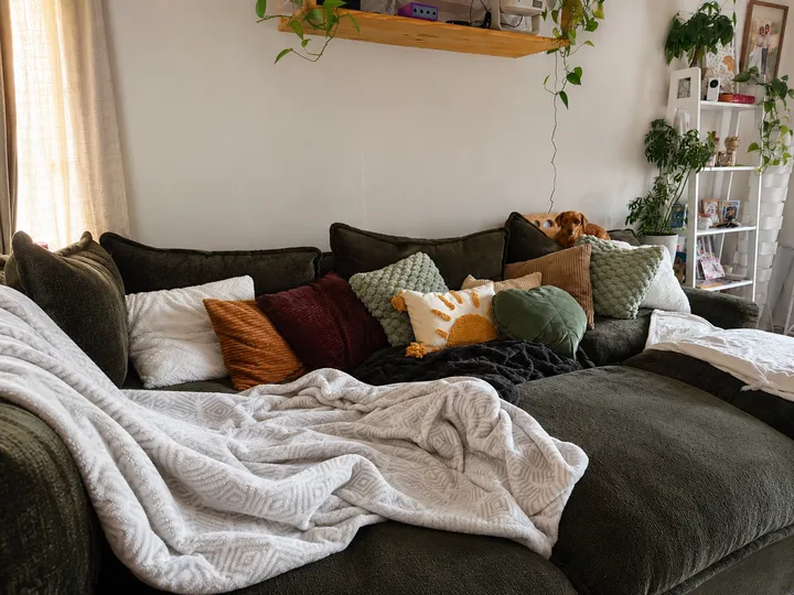 A cozy living room scene featuring a green sofa adorned with various colorful pillows and a soft blanket, creating a warm and inviting atmosphere.