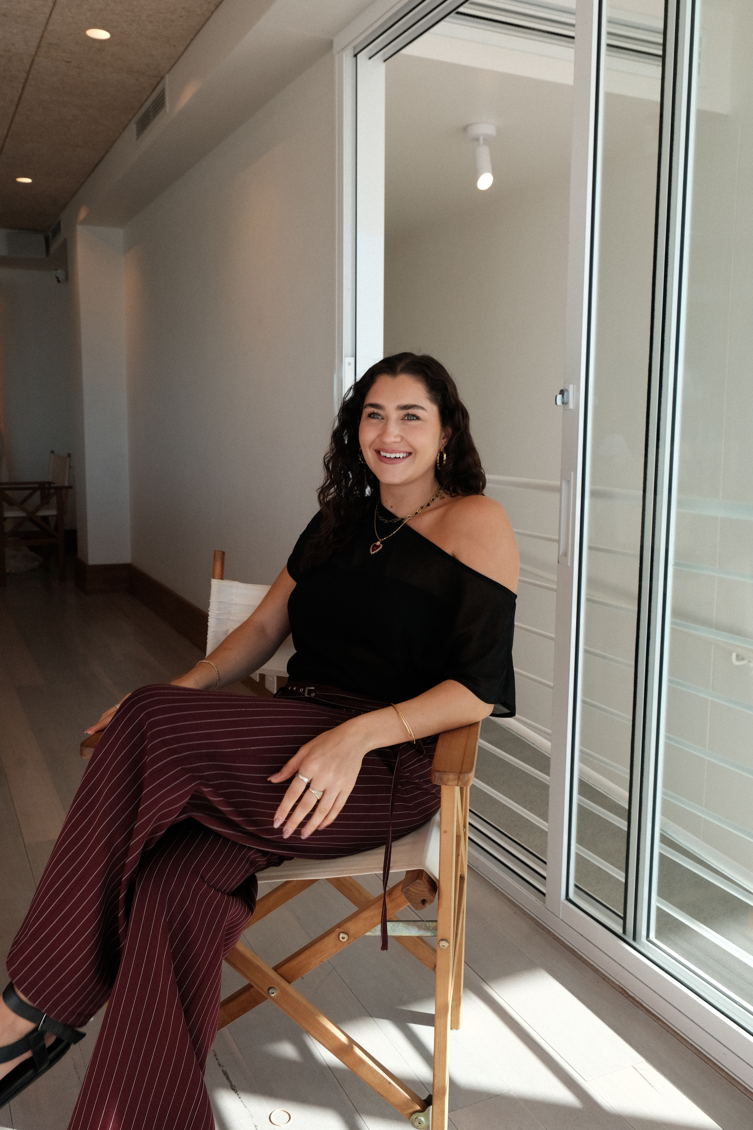 A woman with curly hair sits in a wooden chair, wearing a black off-shoulder top and striped pants, in a bright, modern interior.
