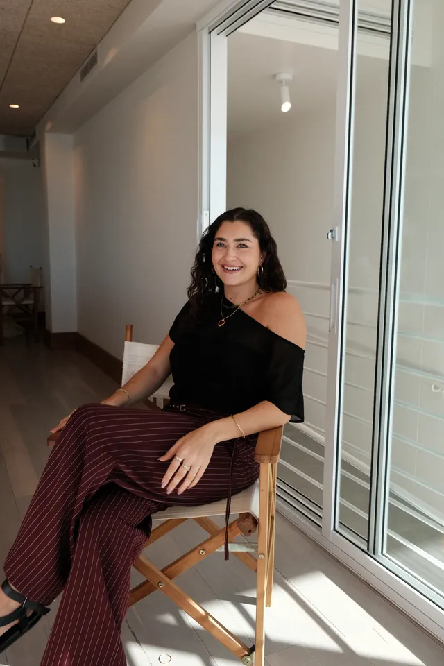 A woman with curly hair sits in a wooden chair, wearing a black off-shoulder top and striped pants, in a bright, modern interior.