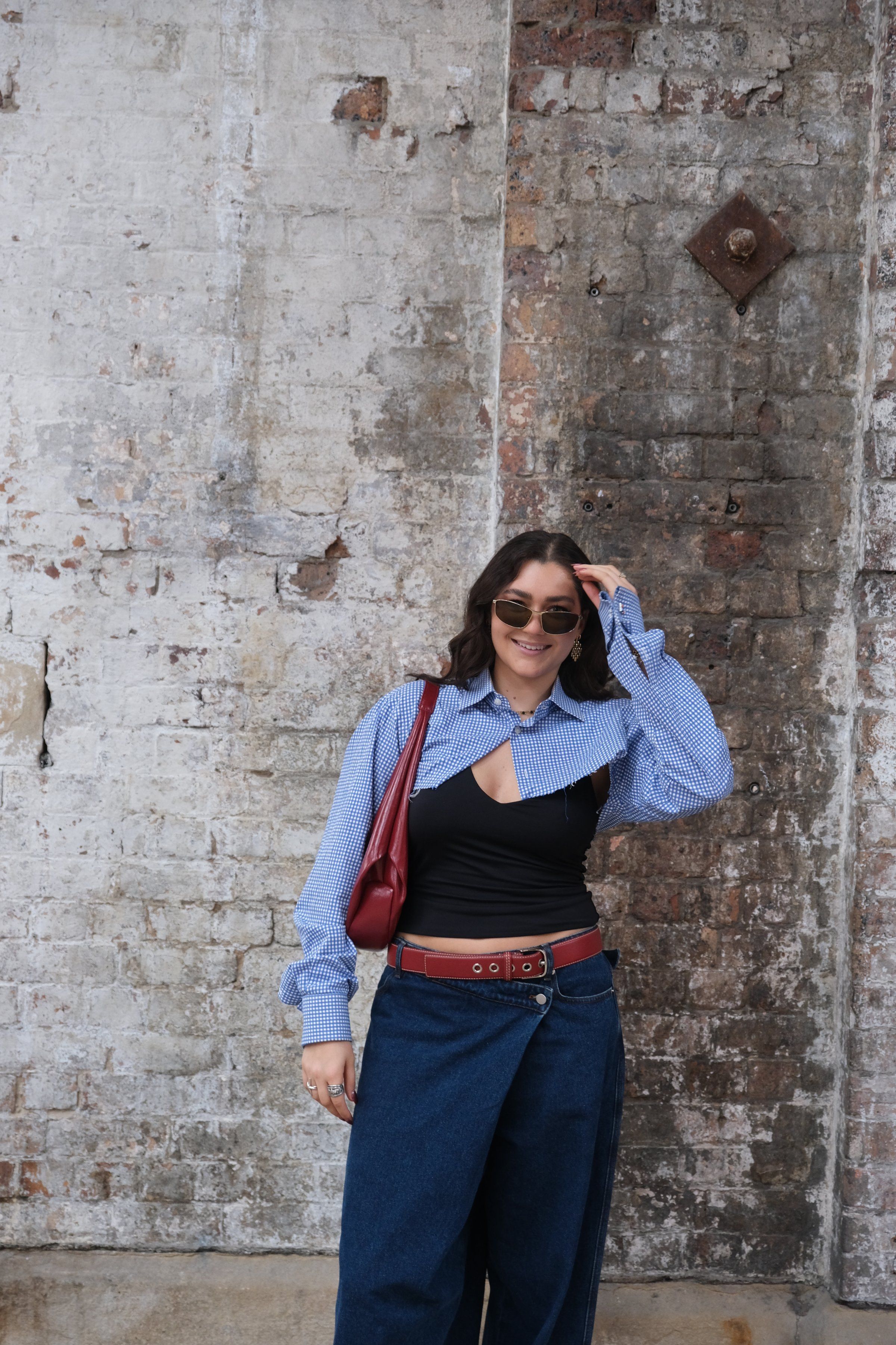 A woman stands against a textured brick wall, wearing a blue and white patterned shirt, a black top, and wide-legged denim pants, accessorized with sunglasses and a red handbag.