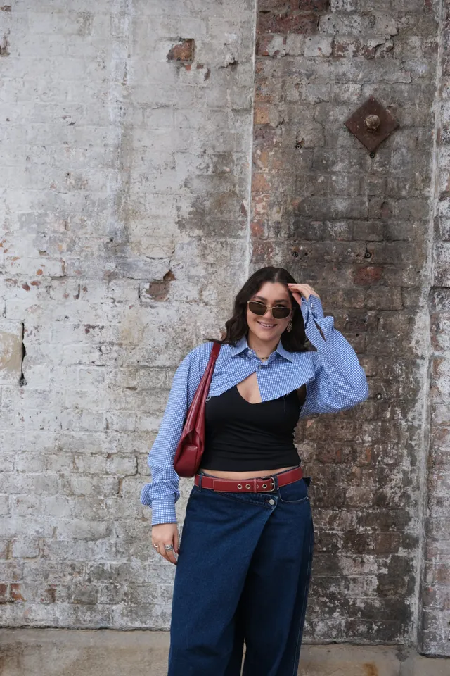 A woman stands against a textured brick wall, wearing a blue and white patterned shirt, a black top, and wide-legged denim pants, accessorized with sunglasses and a red handbag.