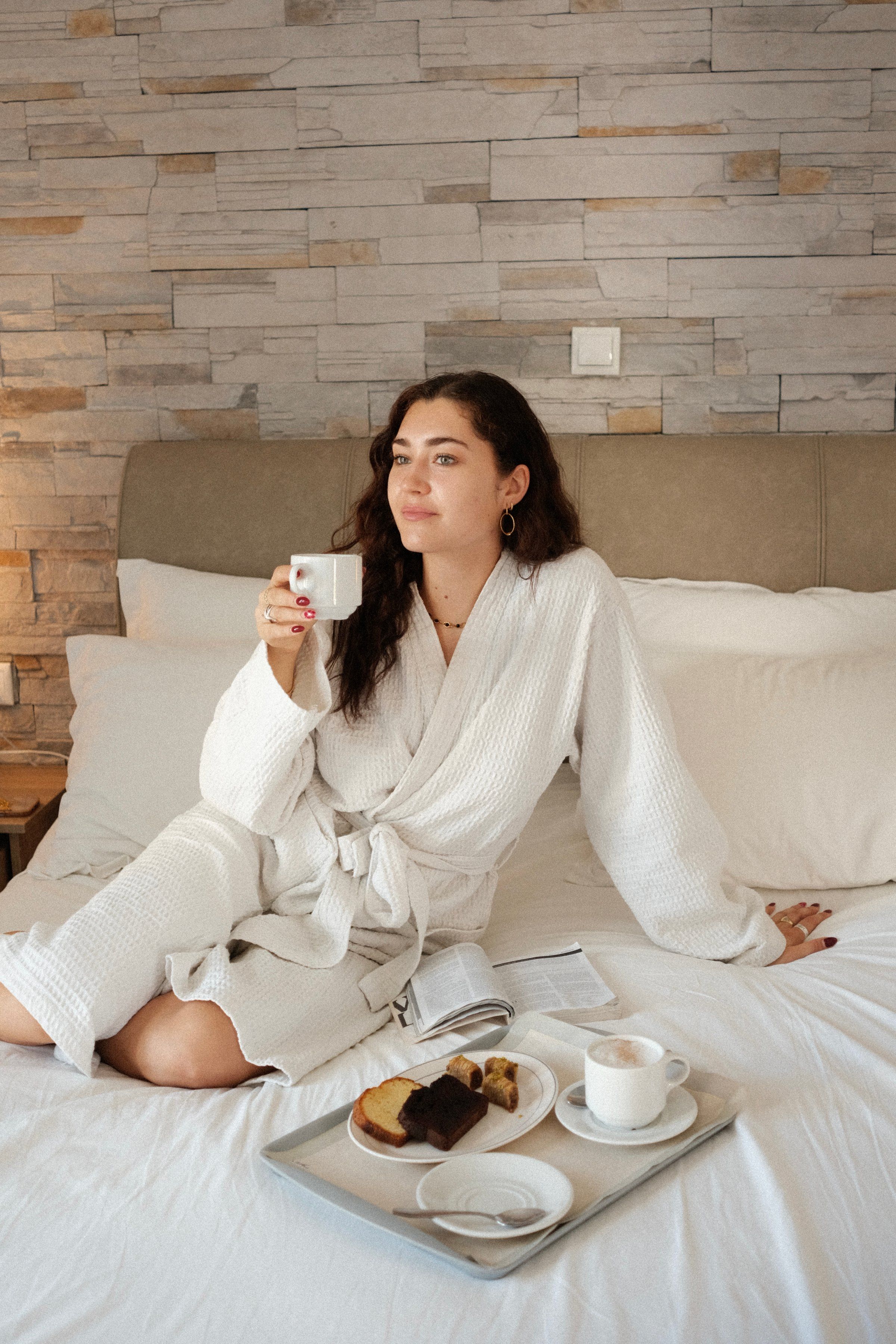 A woman in a white robe sits on a bed, enjoying a cup of coffee with a breakfast tray featuring pastries and a drink beside her.