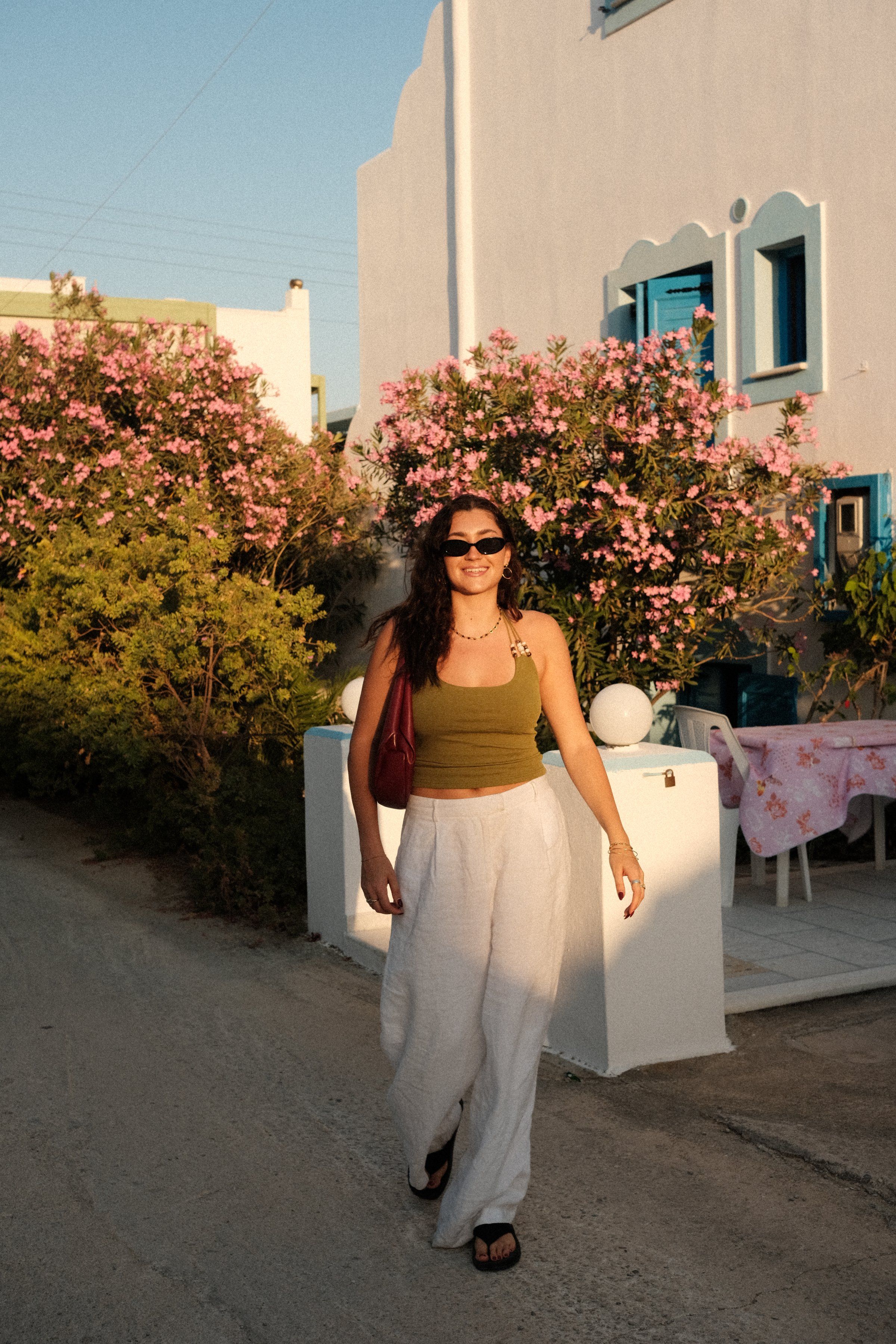 A woman walks along a sunlit path surrounded by blooming flowers, wearing a green top and white pants.
