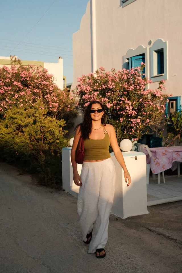 A woman walks along a sunlit path surrounded by blooming flowers, wearing a green top and white pants.
