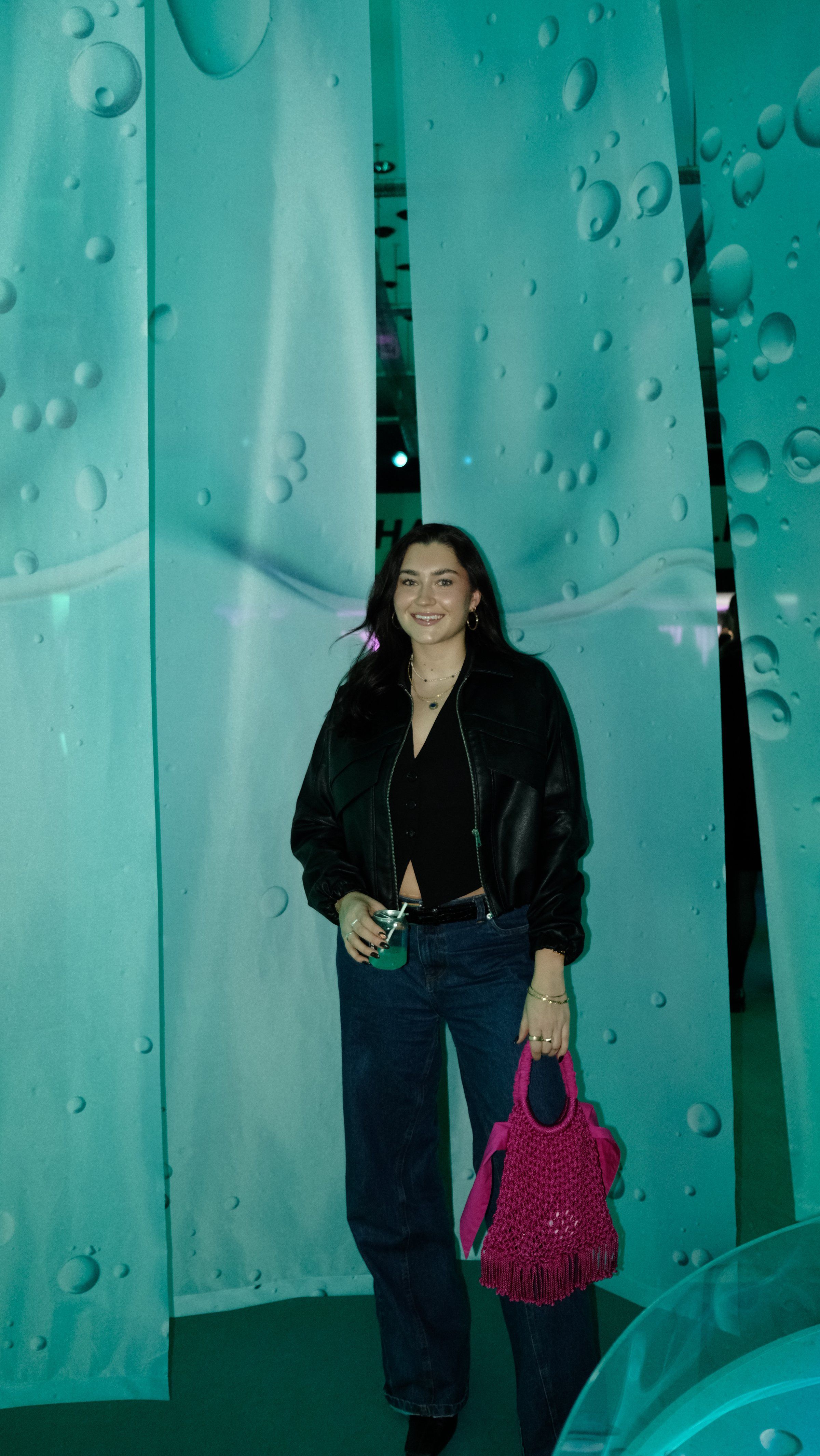 A woman stands confidently in front of a backdrop with a turquoise color and bubble-like patterns, wearing a black top and leather jacket, holding a pink handbag.