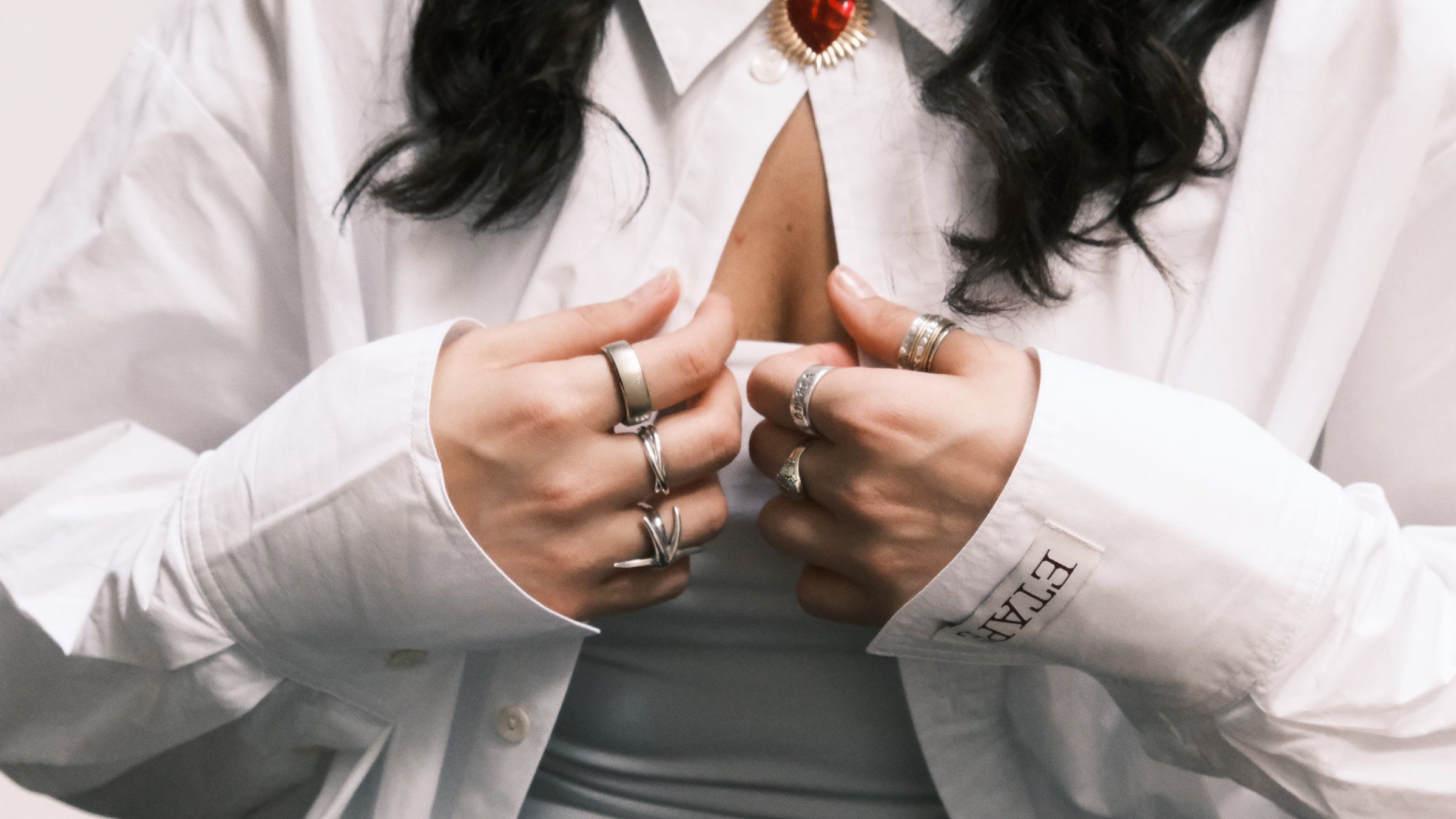 A close-up of hands adorned with multiple rings, holding the collar of a white shirt with a heart-shaped pendant visible.