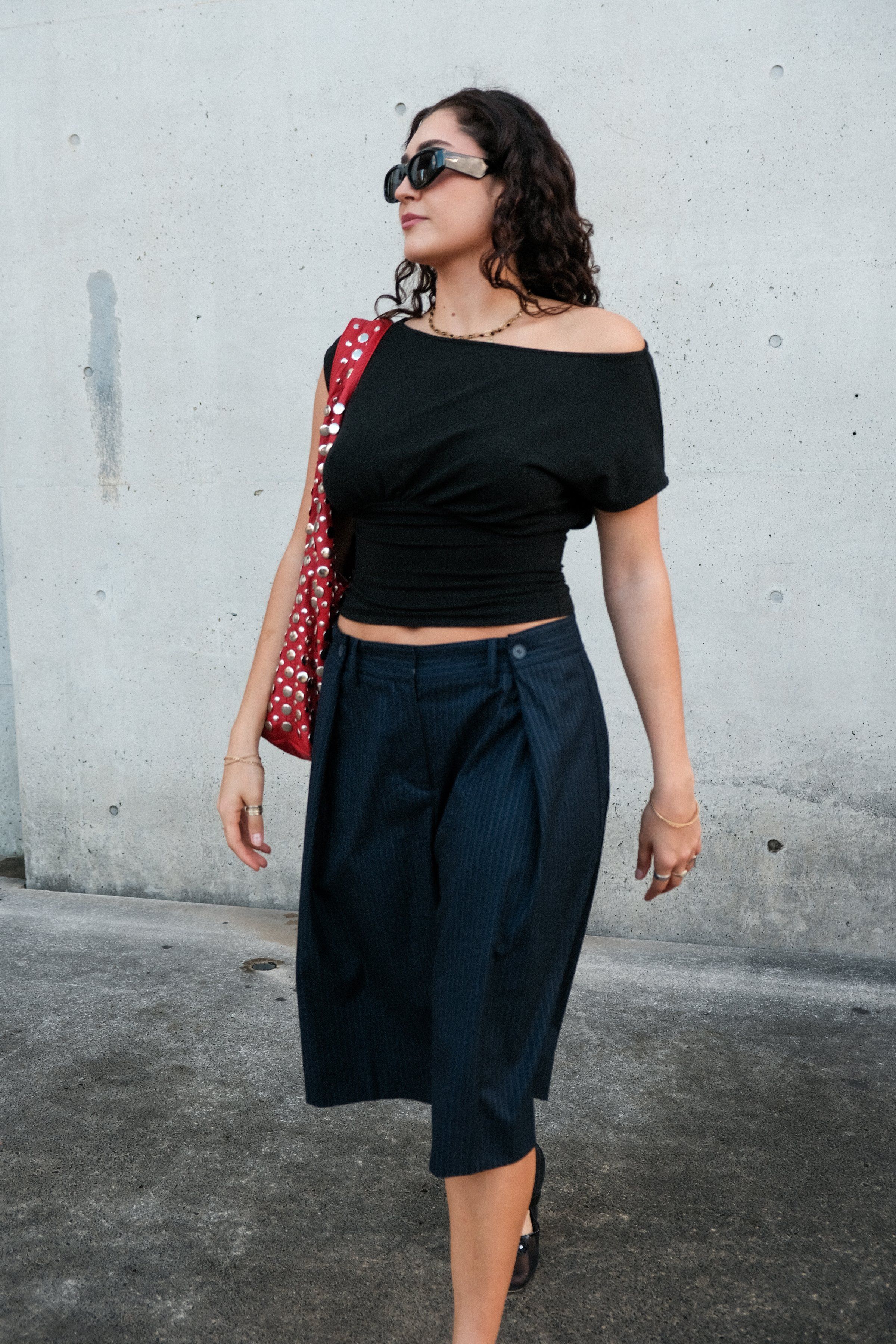 A woman walks confidently in a stylish outfit featuring a black off-shoulder top and navy culottes, accessorized with a red bag and sunglasses.