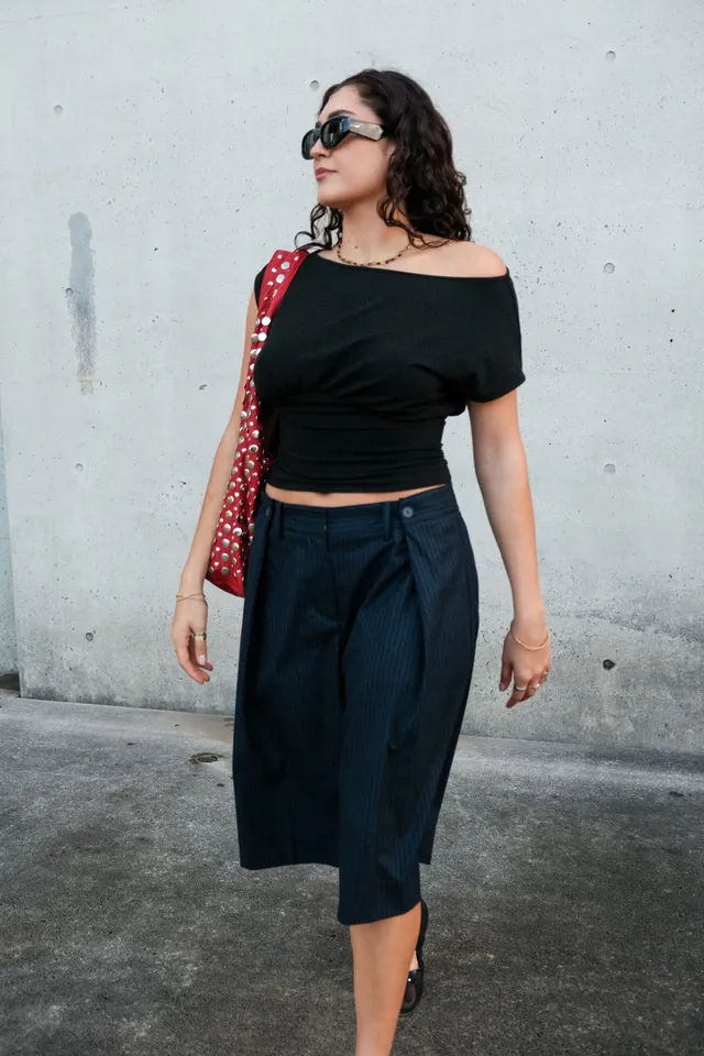 A woman walks confidently in a stylish outfit featuring a black off-shoulder top and navy culottes, accessorized with a red bag and sunglasses.