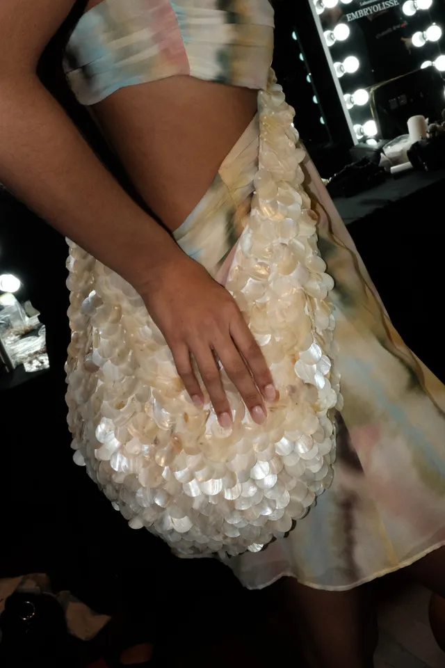 A close-up of a model's hand holding a textured, iridescent bag while wearing a colorful, tie-dye outfit.
