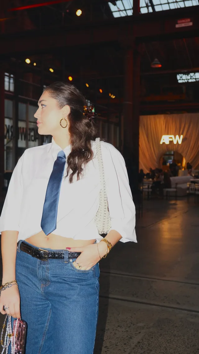 A woman stands confidently in a stylish outfit featuring a cropped white shirt and a tie, paired with high-waisted jeans. The background shows an event space with soft lighting and the letters 'AFW' illuminated.