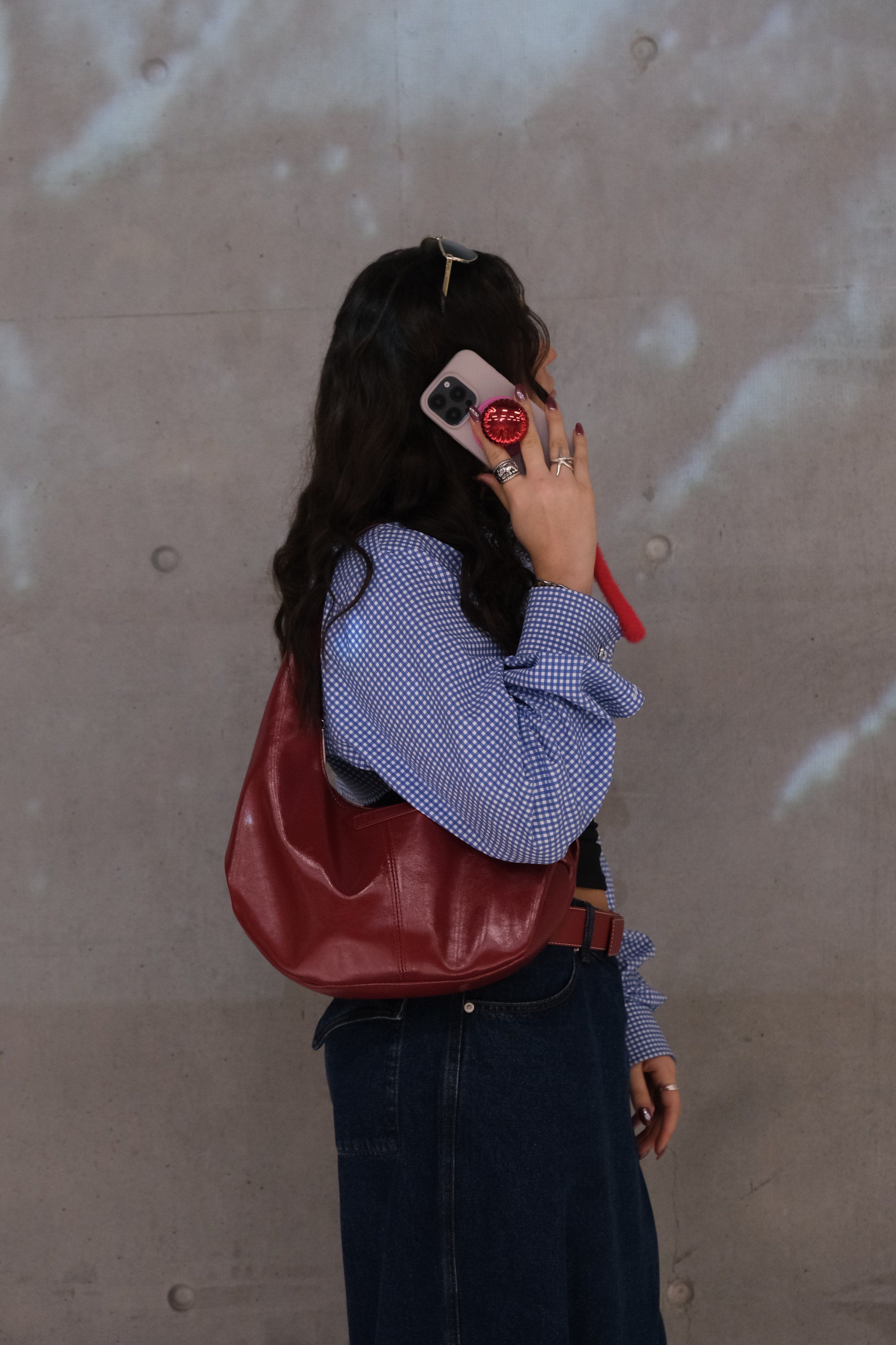 A woman with long, curly hair is holding a phone to her ear while wearing a blue checkered shirt and a red handbag. The background features a textured gray wall.