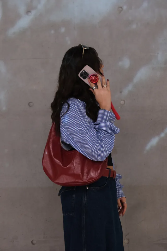A woman with long, curly hair is holding a phone to her ear while wearing a blue checkered shirt and a red handbag. The background features a textured gray wall.