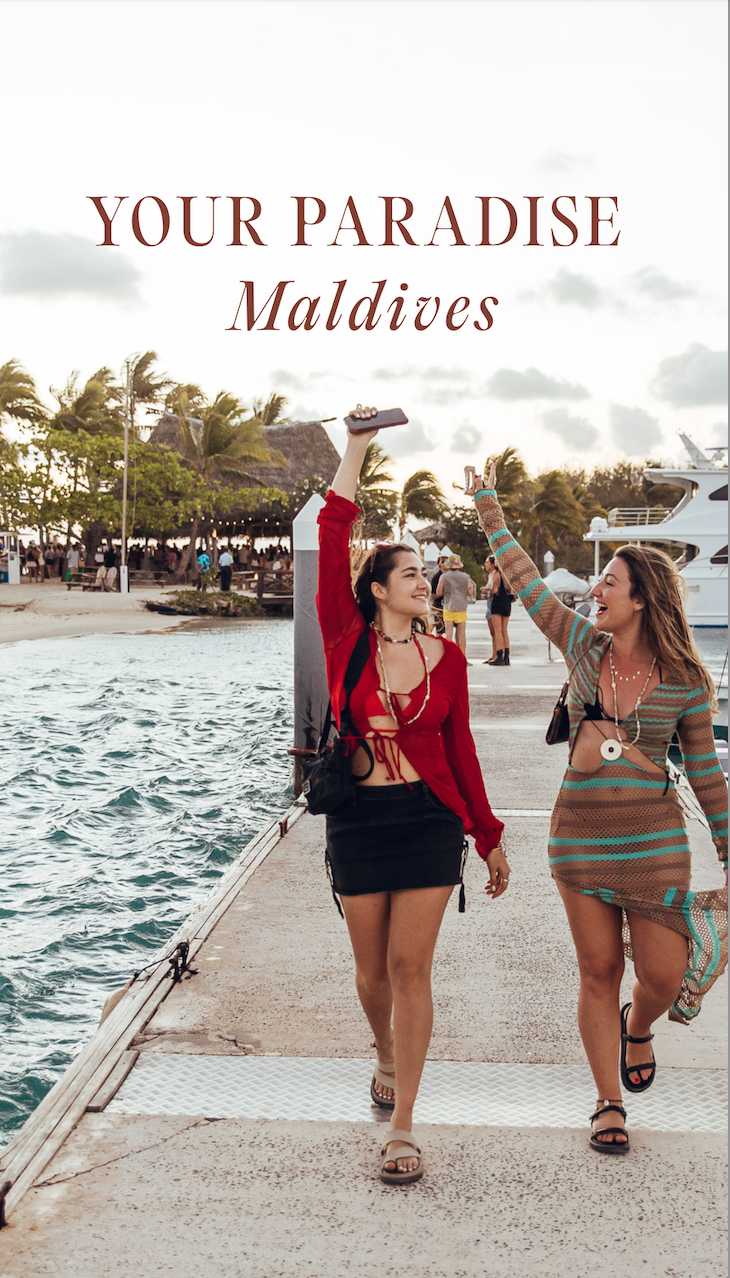 Two women joyfully walking along a dock in the Maldives, holding up their phones. The scene captures a vibrant tropical atmosphere with palm trees in the background.
