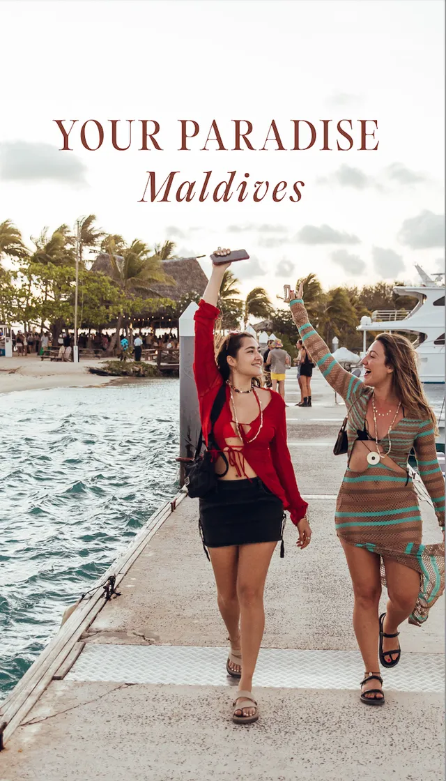 Two women joyfully walking along a dock in the Maldives, holding up their phones. The scene captures a vibrant tropical atmosphere with palm trees in the background.