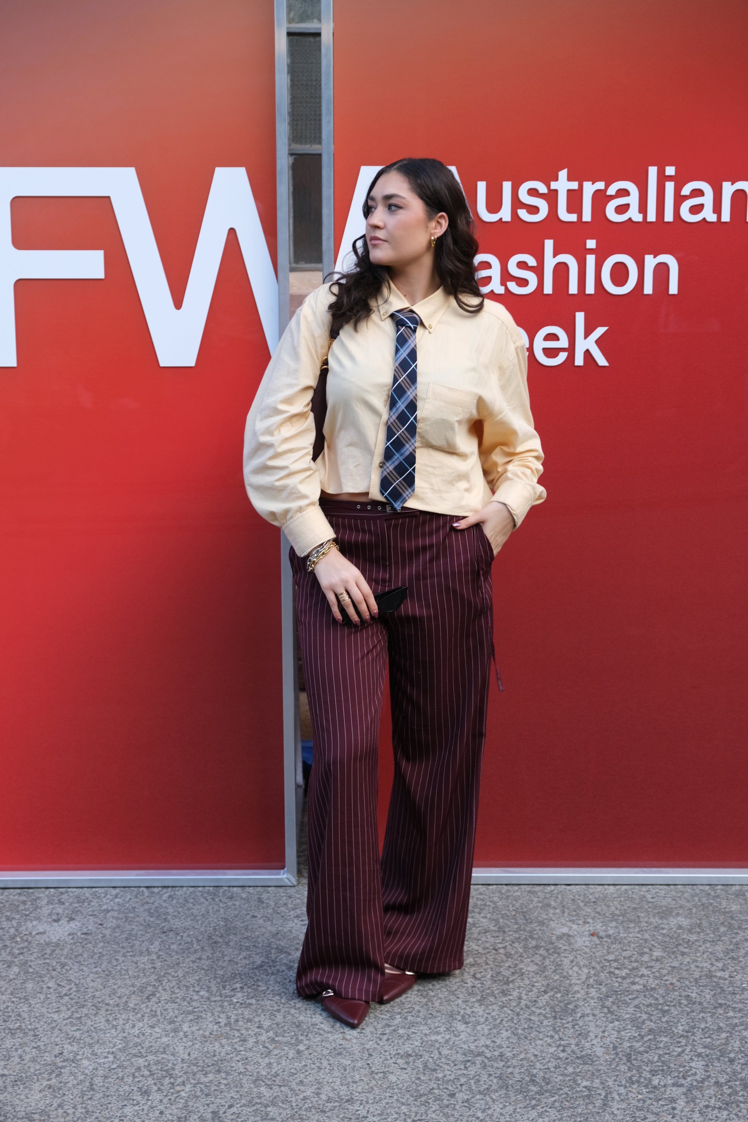 A model poses confidently in a stylish outfit featuring a yellow shirt and a tie, paired with maroon pinstriped pants, against a vibrant red backdrop for Australian Fashion Week.