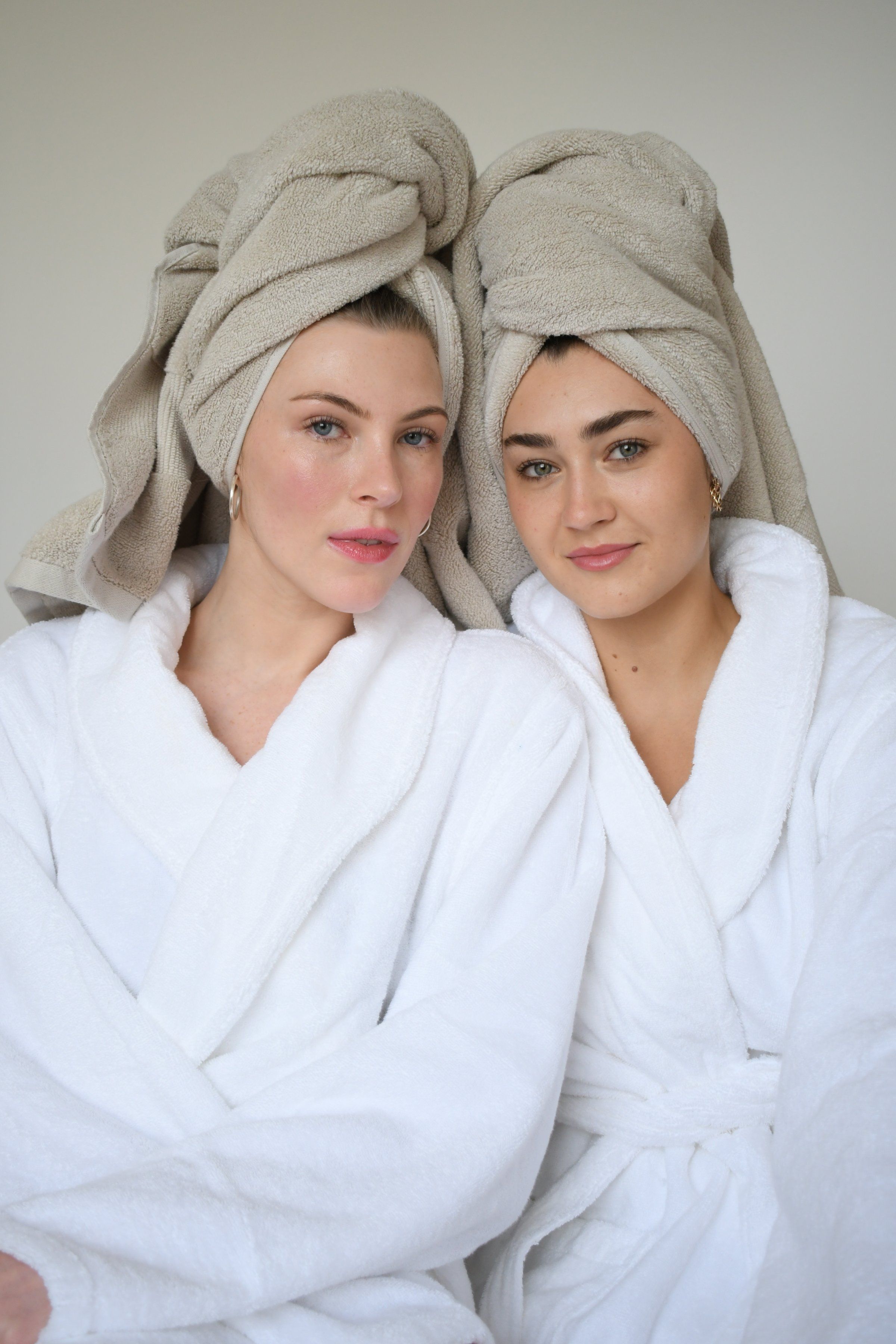 Two women with towels on their heads, wearing white bathrobes, posing together in a relaxed setting.