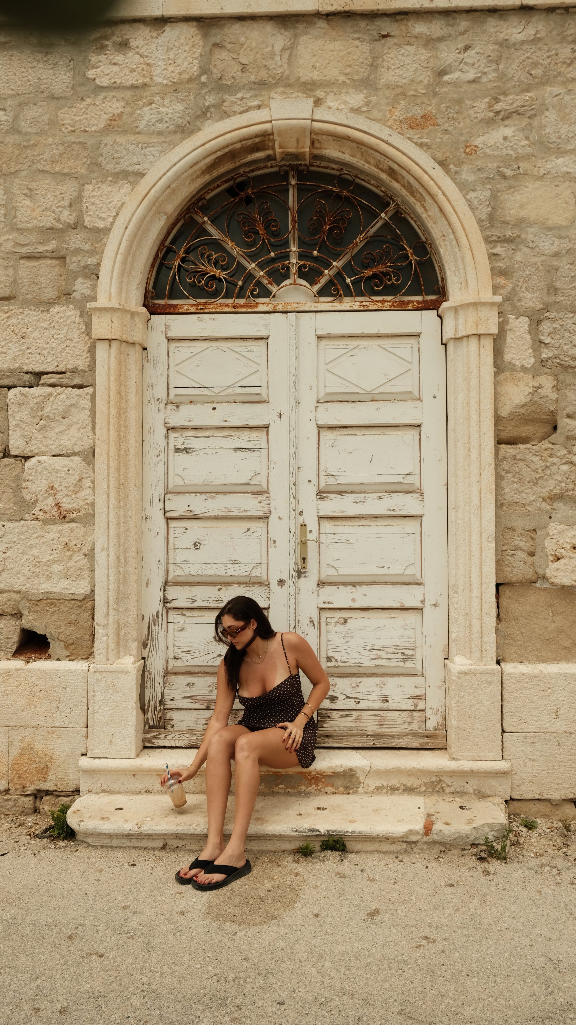 A woman sitting on the steps of an old stone building, wearing a patterned dress and sunglasses, holding a drink.