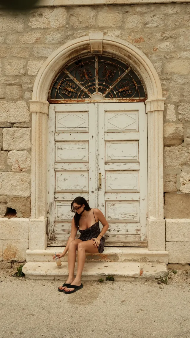 A woman sitting on the steps of an old stone building, wearing a patterned dress and sunglasses, holding a drink.