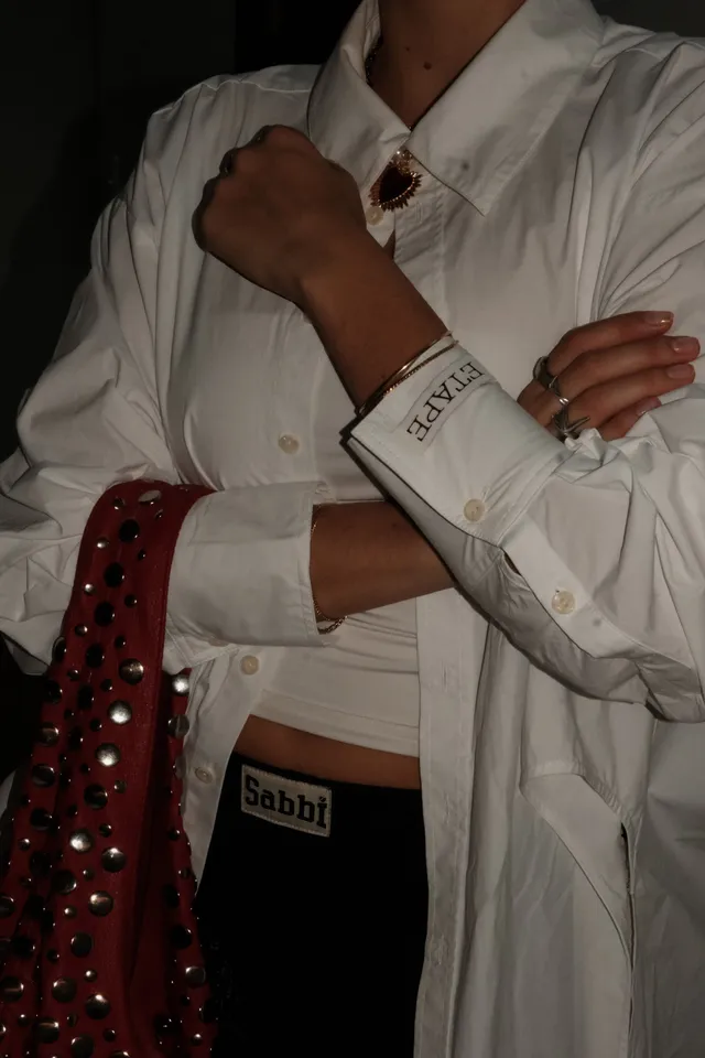 A close-up of a person wearing a white oversized shirt with a cropped top underneath, accessorized with jewelry and holding a red bag adorned with studs.