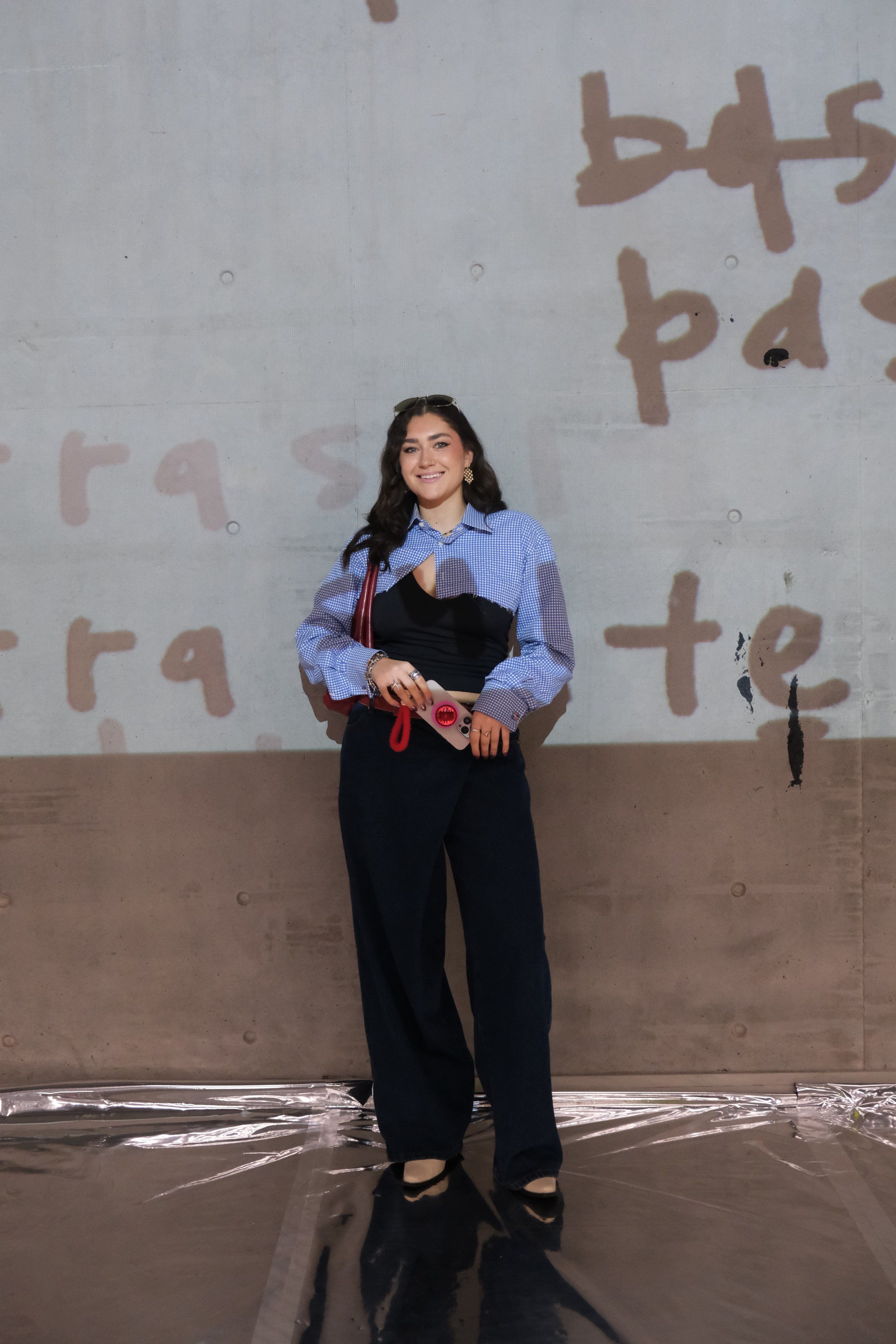 A woman stands confidently in front of a textured wall, wearing a stylish outfit that combines a patterned blouse and tailored pants, holding a pair of red scissors.