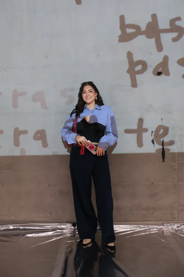 A woman stands confidently in front of a textured wall, wearing a stylish outfit that combines a patterned blouse and tailored pants, holding a pair of red scissors.