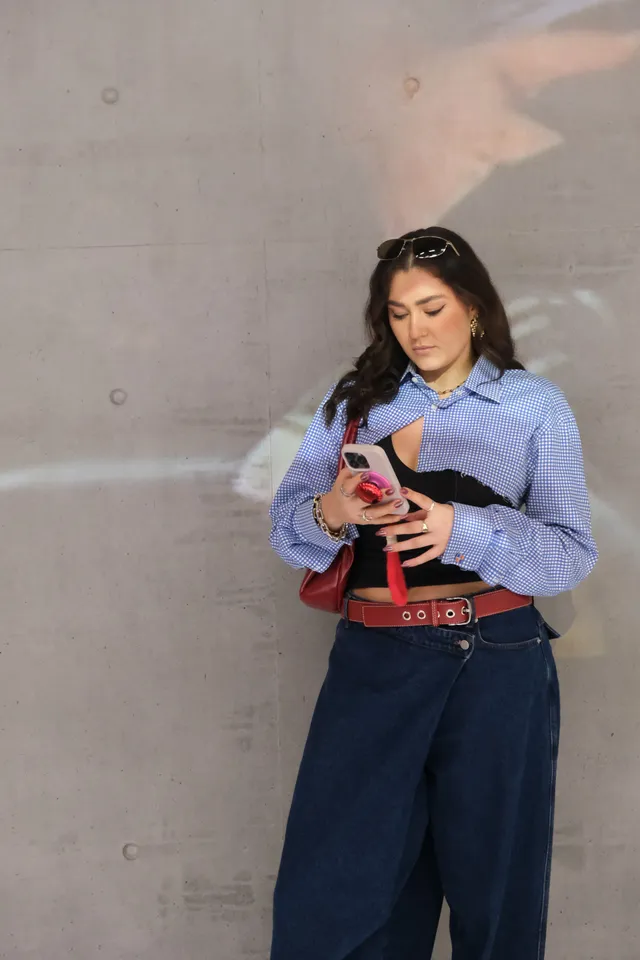 A woman stands against a concrete wall, looking at her phone while wearing a stylish outfit that includes a checkered shirt and wide-leg jeans.
