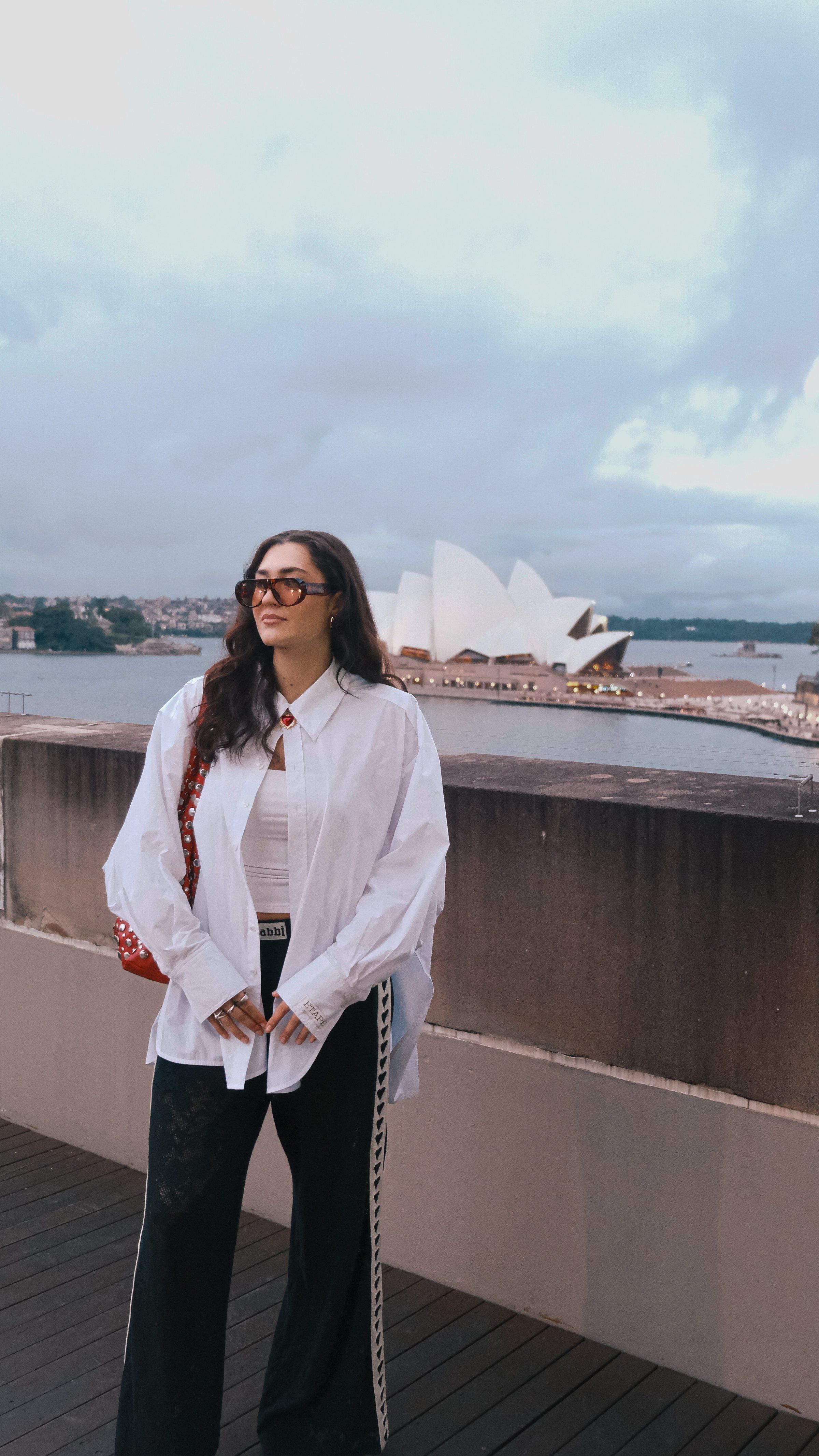 A woman stands on a rooftop terrace with the Sydney Opera House in the background, wearing a white oversized shirt and black pants.