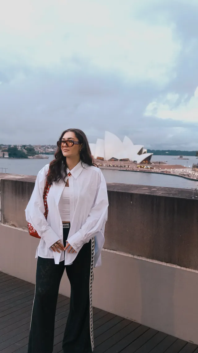 A woman stands on a rooftop terrace with the Sydney Opera House in the background, wearing a white oversized shirt and black pants.