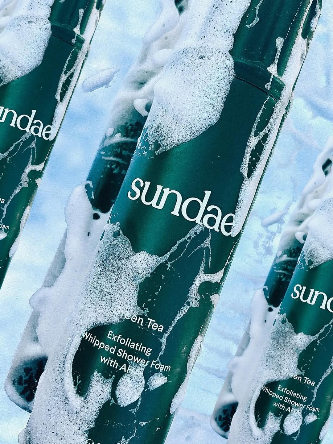 A close-up of a bottle of exfoliating whipped shower foam labeled 'sundae' with a green tea scent, surrounded by bubbles.