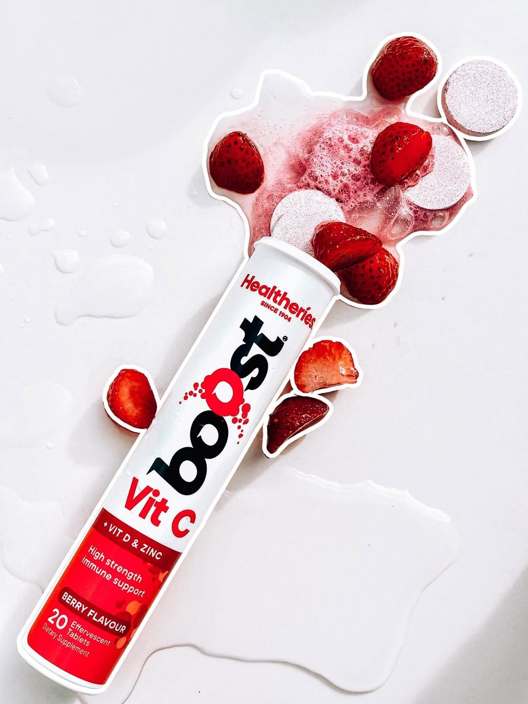 A vibrant product shot of a Healthieries effervescent vitamin C tablet tube surrounded by fresh strawberries and splashes of berry flavor.