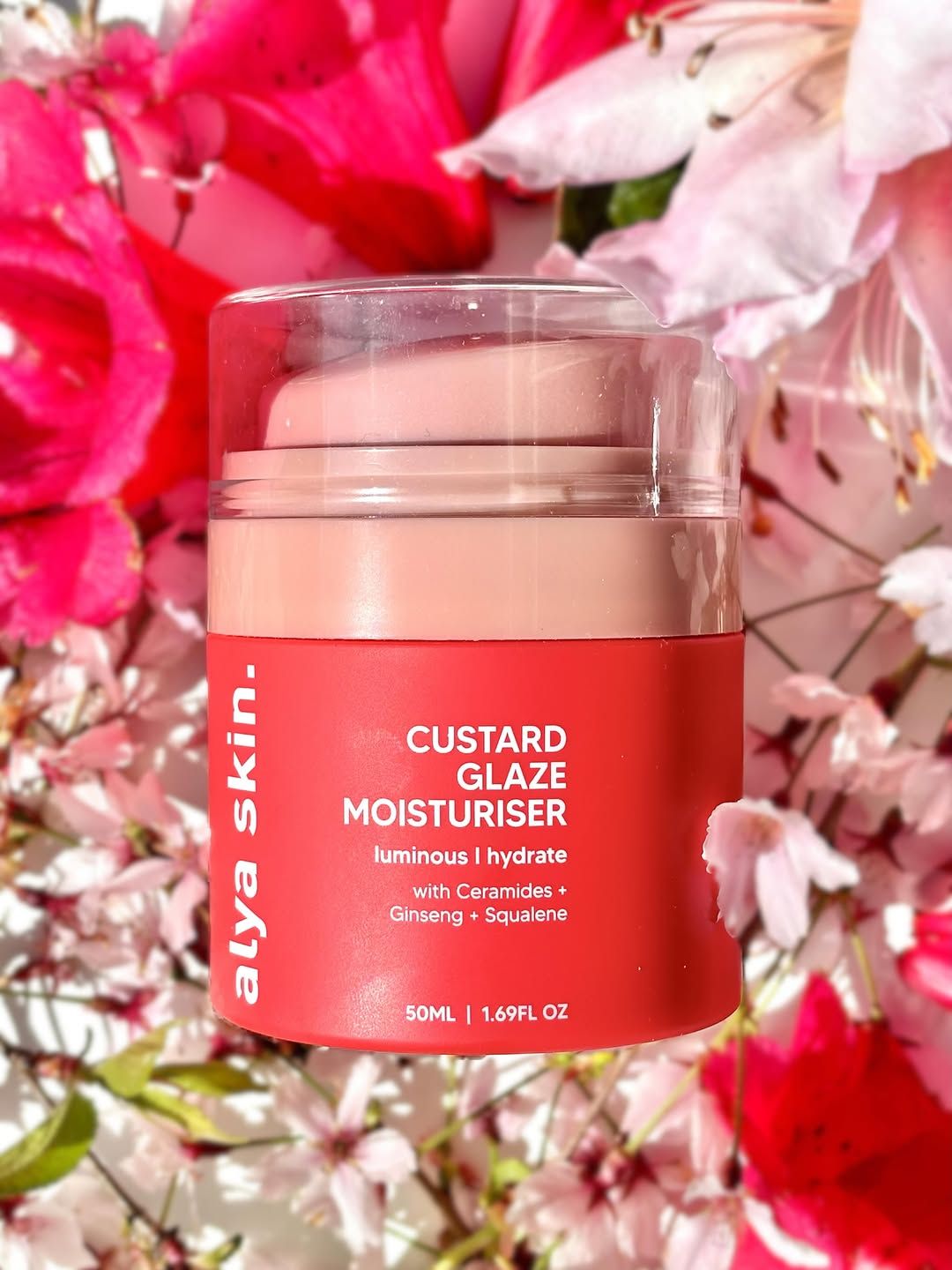 A jar of Alya Skin Custard Glaze Moisturiser surrounded by pink flowers, showcasing its vibrant packaging.