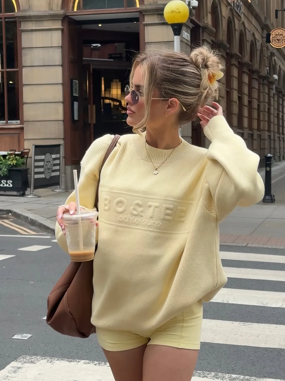 A woman in a light yellow oversized sweater and shorts holds a drink while walking on a city street. She has sunglasses on and her hair is styled in a casual updo.