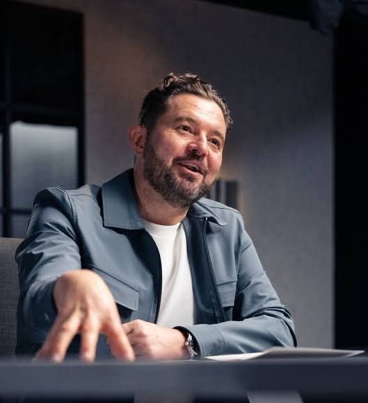 A man with a beard and curly hair is seated at a table, gesturing while speaking in a modern, well-lit room.